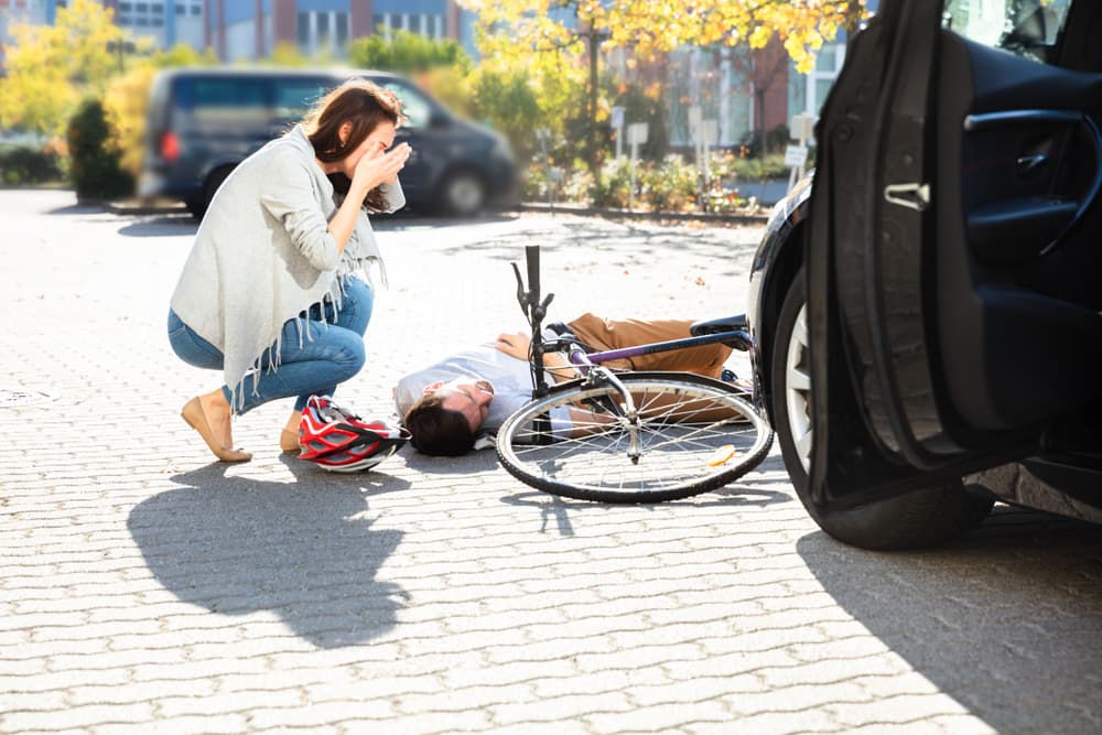 Woman Looking At Unconscious Male Cyclist Lying On Street After Accident Near Car