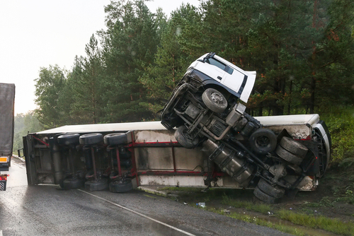 A semi-truck overturned on the side of a road in a forested area, with its trailer lying on its side.