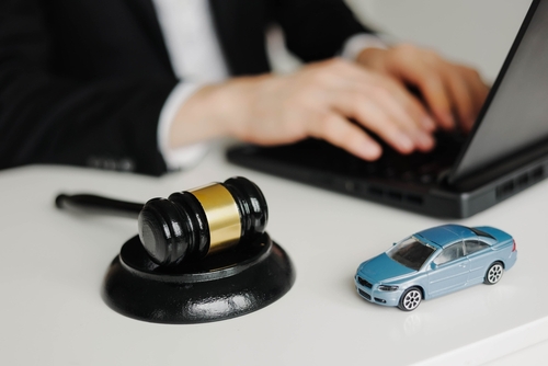 A workspace with a gavel and a toy car on a desk, while a professional works on a laptop in the background, indicating legal research or case preparation for a car accident claim.