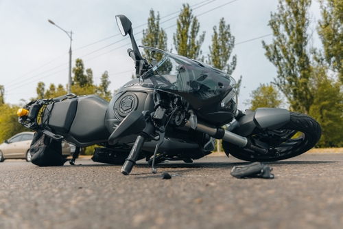 A black motorcycle lying on its side on an asphalt road, with debris scattered nearby and trees in the background.