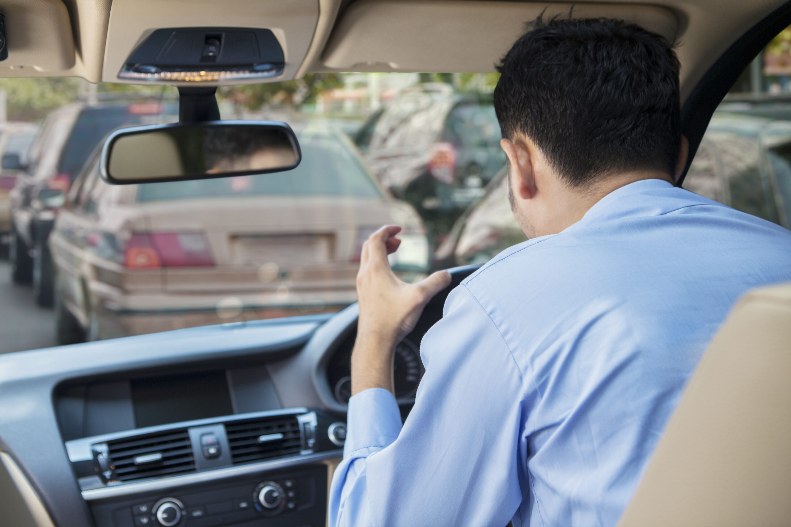 A man seated in his car, visibly frustrated, with his hand raised in an agitated gesture. The traffic jam outside the vehicle suggests a typical situation where road rage may arise.