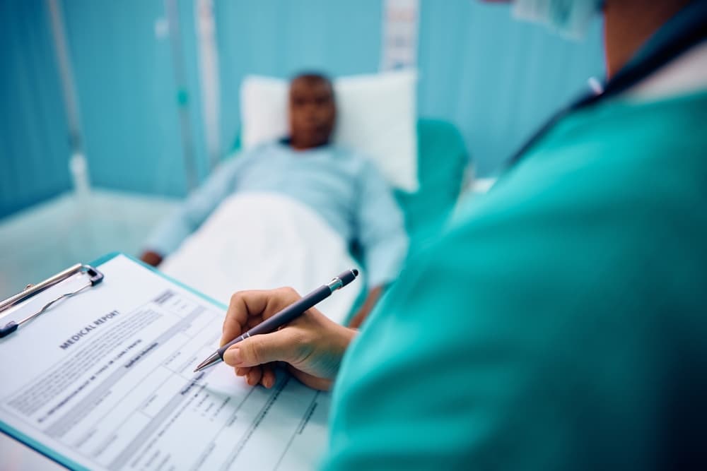 A medical professional in green scrubs filling out a medical report on a clipboard while a patient lies in a hospital bed in the background.