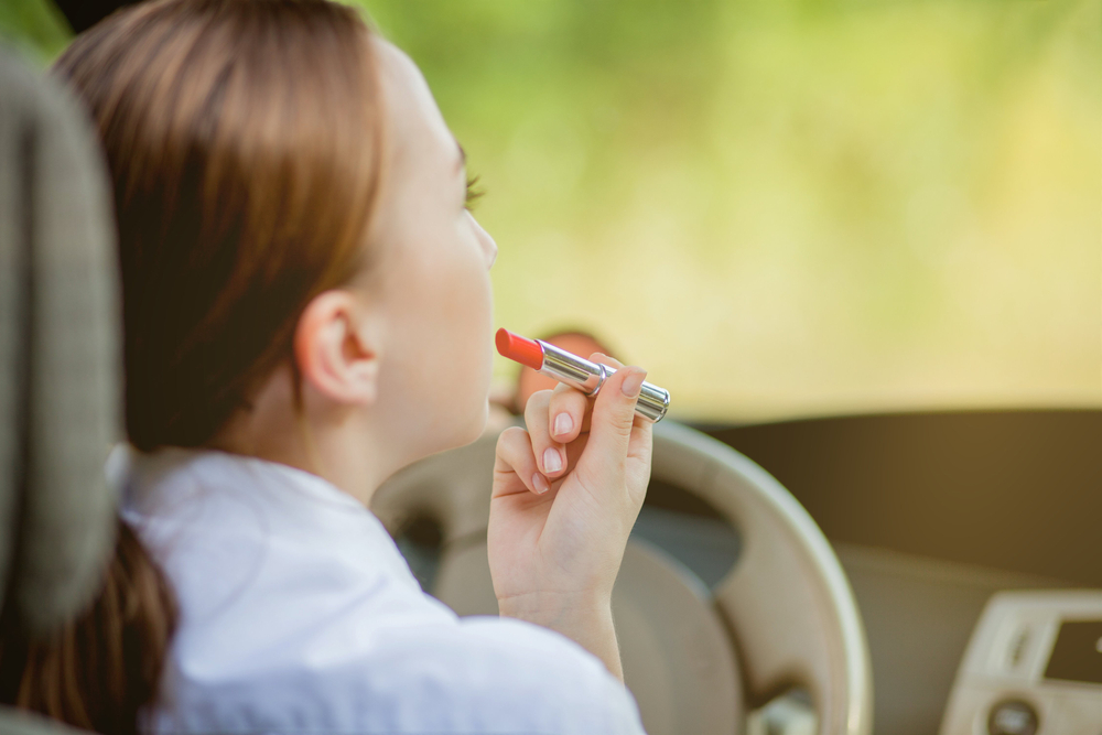 Young woman driver red haired teenage girl painting her lips doing applying make up while driving the car.